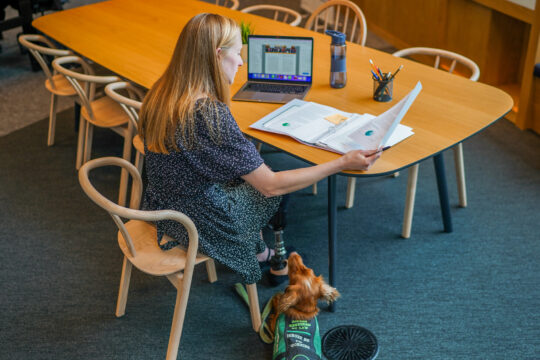 A woman with an assistance dog, and a prosthetic leg, sitting at a table working.