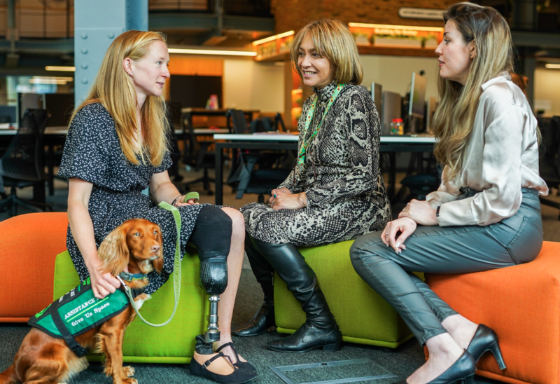 Three (3) women sitting in a colourful breakout area, one of the women has a prosthetic leg and her assistance dog is sitting beside her. One of the women is wearing a sunflower lanyard.